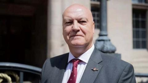 Ian Cooper, a man wearing a dark grey suit and a dark red tie, is standing outside Stafford County Buildings. He is also wearing a white shirt and a pin badge on his lapel.