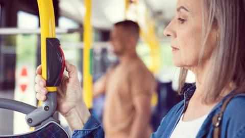 A woman presses the red stop button as she stands on a bus. A man can be seen standing in the background.