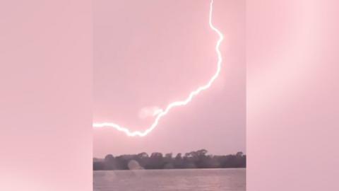 Image of a large arc of lightning streaking across the sky. The bolt is bright enough to tinge the sky a reddish pink.
