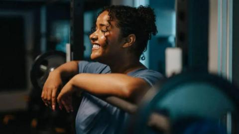A woman with curly brown hair is in a gym, leaning on a weight. She is wearing a grey t-shirt and smiling