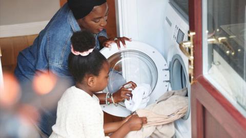 A mother and daughter put laundry into a washing machine together in their home