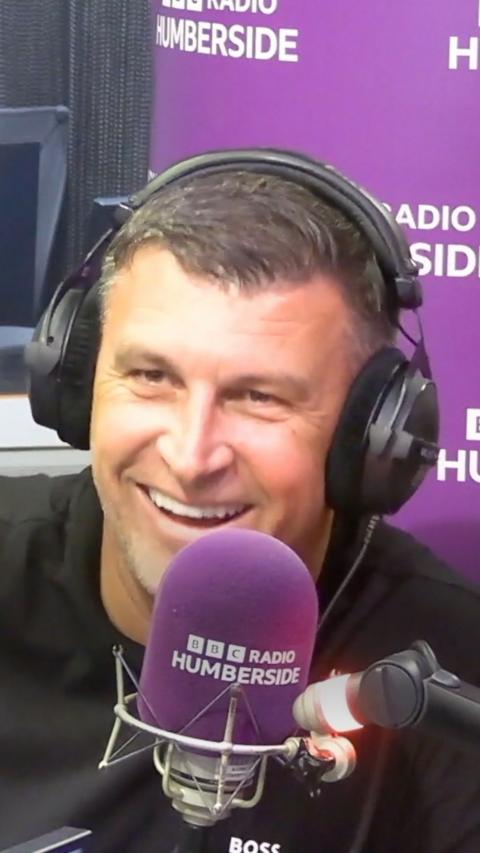 A man wearing headphones smiling behind a purple microphone.