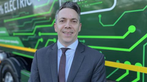 Cabinet member for waste and environment councillor Dominic Muns smiling in a shirt and tie in front of the new electric bin lorry.