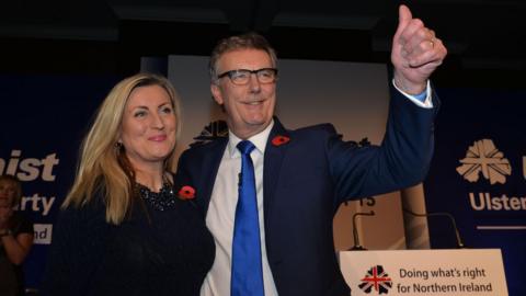 Linda Bryans wears a black top and is smiling, looking into the distance. She has blonde hair and wears a poppy. Mike Nesbitt is wearing a blue suit and blue tie and poppy. He has grey hair and wears glasses. He is doing a thumbs up sign. There are UUP banners behind.