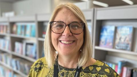 Kerry Murray, a woman who is standing inside a library in front of a grey book shelf which has several books on it. She has shoulder length blonde hair and is wearing a yellow and black T-shirt and a black lanyard. She is looking directly at the camera and smiling and has a pair of glasses over her eyes.
