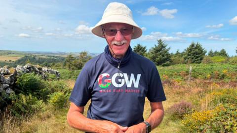 John Parry stands smiling at the camera, with a view of north Pembrokeshire and the Pembrokeshire coast behind him. John is wearing a 'grandad's great walk' fundraiser logo t-shirt, with sunglasses and a sun hat. He wears sunglasses and a white moustache.