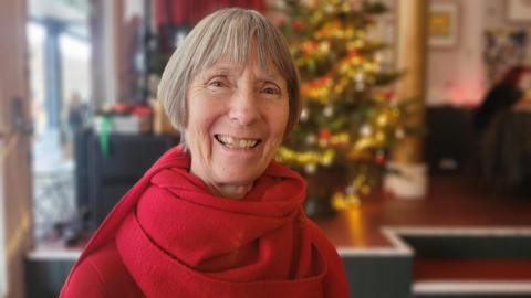 Alexandra Pickford is sitting in a cafe dressed in a red top and red scarf. She has short, grey hair and is smiling. There is a Christmas tree lit up behind her.