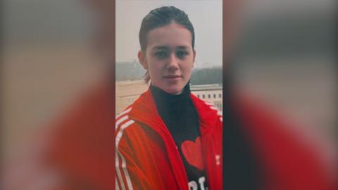 A girl with pulled back brown hair  is wearing a high-necked black top with red and white decoration on it, and a red jacket.