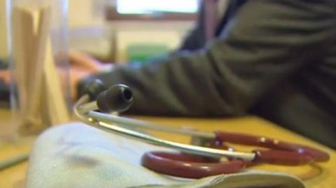 A stethoscope on a table in the fore, with a blurred background of someone in dark clothing sitting at the table, with arms resting on it.