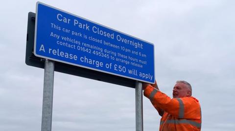 A large blue metal sign is being secured by a man in orange hi-vis at the problem car park. The sign reads: Car Park Closed Overnight. This car park is closed between 10pm and 6am. Any vehicles remaining during these hours must contact 01642495345 to arrange release. A release charge of £50 will apply.