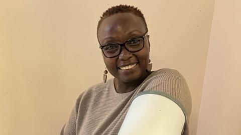 A person wearing a beige jumper and glasses has their arm in a white metal cylinder which is testing their blood pressure