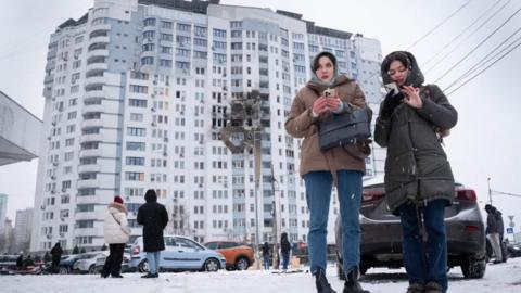 Two women stand wrapped up in winter clothing. There is snow on the ground and the building behind them has a large dark scorch mark where it has been hit with an explosive 