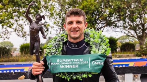 Lewis Arrowsmith, who is wearing black leathers and has a laurel garland around his neck with a green banner that says Supertwin Manx Grand Prix Winner across it. He is holding a bronze MGP trophy.