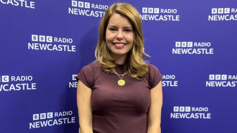 Kim McGuinness smiling into the camera in front of a purple BBC Radio Newcastle backdrop. She has curled, blonde hair which falls just below her shoulders and is wearing a large gold necklace and a purple t-shirt. 