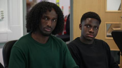 Adnan, 18, wears a dark green top and is sitting at a table inside an office room beside Edem, 18, in a black top, both listening to the discussion 