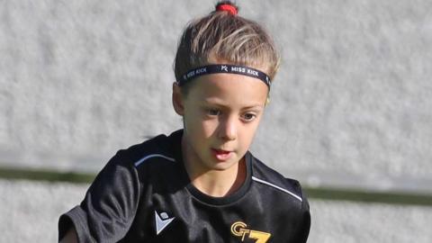 Poppy Atkinson playing football. Her hair is pulled back into a ponytail and she is wearing a black football kit. She is looking down at the ground.