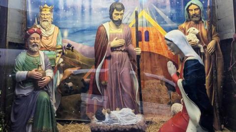 A nativity scene of life-size figures behind a pane of glass. To the left are two kings, dressed in green, purple and peach robes. The one that is standing wears a crown and the one kneeling wears a turban. In the centre, a baby lies in white swaddling in a manager. Standing over him is a man dressed in red robes holding a long stick. To the right, a woman is kneeling in prayer dressed in blue, red and a white headdress. Behind her is a shepherd in brown robe, holding a sheep in one hand and a long walking stick in the other. They all stand on a bed of straw with a printed backdrop of a stable on a starry night.