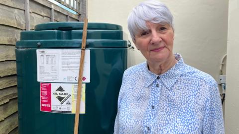 A woman wearing a blue and white blouse stands in front of a green heating oil tank.
