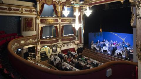 A view from a theatre balcony looking over a group of people on table's looking up at the opera house's stage