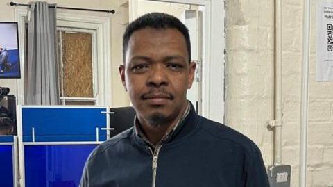 A man stands in front of the entrance to an office indoors. He has short-cropped black hair and a moustache and goatee. 