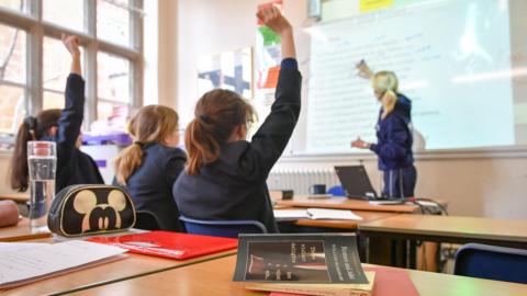 Three children in a classroom - two with their hands raised. A teacher is writing on a whiteboard. Everyone is dressed in blue and the children are wearing blue jackets. In the foreground there are two books on a table.