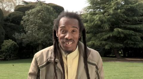 The late poet Benjamin Zephaniah, wearing a yellow shirt and beige jacket. He is standing outside, in an open park area with trees behind.