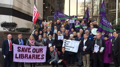 A large group of people standing in front of a building. Some of the people are holding flags and banners that read 'save our libraries' 