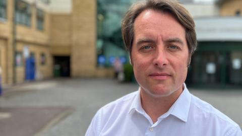 A head and shoulders picture of Thom Lafferty. He is standing in front of a hospital and is wearing a white shirt with the top button undone.