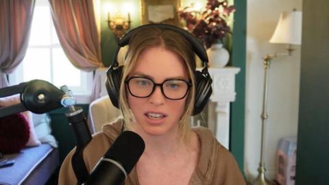 A woman, QTCinderella, is sitting wearing black over-ear headphones and black glasses, with a brown, open hoodie. She is speaking into a black microphone. In the background is a lamp and a vase of flowers and a window with pink curtains.