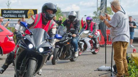 Three motorcyclists in a line setting off with a few people around them watching them go