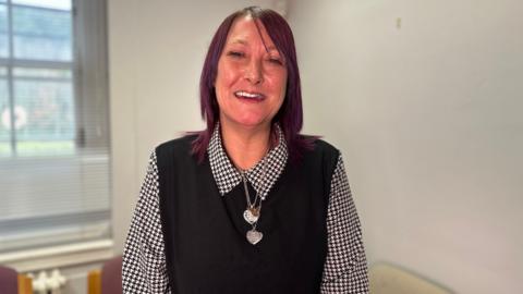 Claire Curran is smiling. Her shoulder length hair is styled in maroon. She's wearing a black vest jumper with a black and white blouse underneath and heart shaped pendants are around her neck. She's standing in a room. 