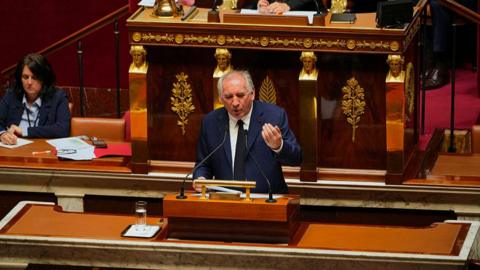 French Prime Minister François Bayrou addresses members of parliament before a non-confidence vote. He is wearing a suit. 