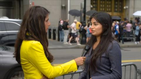 Nomia Iqbal, who has long brown hair and wears a yellow blazer, holds a mic towards a reporter who also has long dark hair and wears a blazer, outside of a New York City courthouse where photographers are waiting