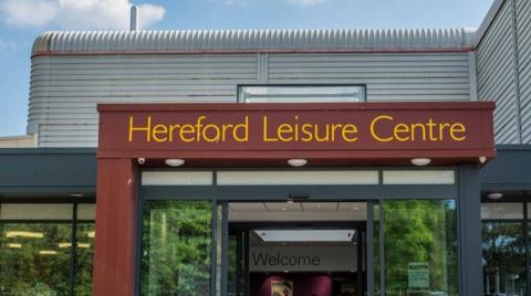 Entrance to Hereford Leisure Centre, with that name being in yellow letters above the door. Blue sky can be seen above the silver building.