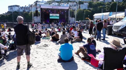 A large festival stage on a beach. Scores of people are sitting in front of it. 