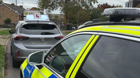 A grey Vauxhall with a white and red learner sign on top of it has pulled over to the side of a residential road. Behind it (and in the foreground) is a police car with the usual yellow and blue branding.