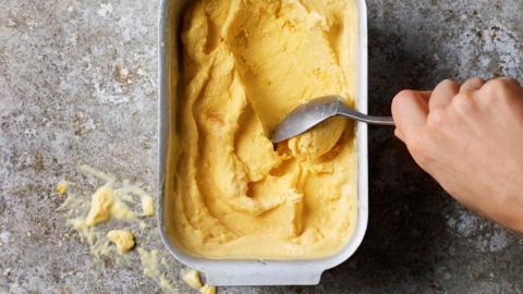 A person scoops yellow ice cream out of a container using a spoon