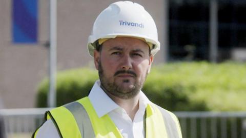 Patrick McIntyre - a man with a thin beard and moustache, with dark hair. He has a high vis jacket and white hard hat on. 
