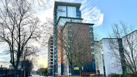 Modern 16-storey block of flats with some bare trees in the foreground.  The building has orange and white cladding with 'Unite Students' written on one side near the top.