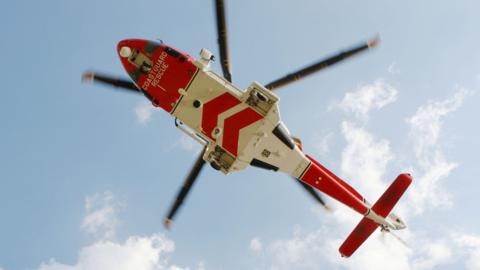 A view from the ground up towards the underside of a coastguard helicopter. It is viewed against a blue sky with clouds. The helicopter has red and white markings.