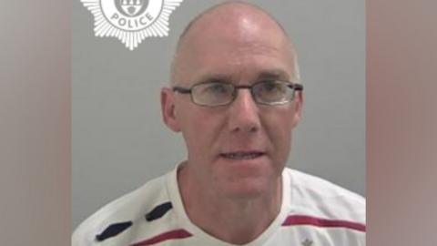 A mugshot of a bald man wearing black glasses, and a white t-shirt with a red and black horizontal stripe.