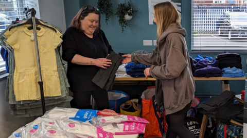 A woman in a black t-shirt is holding some grey children's trousers. A woman to her right, wearing a brown hoodie, is passing the trousers to her. To the left is a clothes rail with coloured checkerboard school dresses hanging on it. There are also tables piled with other clothing.