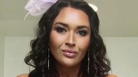 A woman with long dark hair with a pink bow in it and dark eyes looks straight into the camera. 