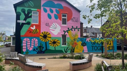 The garden, which was built on an empty plot next to Chillingham Road. It features improved pathways, curved benches, trees, planters for bushes and flowers, insect boxes, and talking tubes - play equipment which allows children to talk to each other at opposite ends of the garden. On the outer wall of a house facing the garden is a brightly coloured mural painted by artist Molly Bland, which features cartoon flowers, insects, and birds. The words "Growing Together" are painted at the bottom.