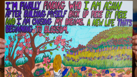 A posted of a painting shows a woman sitting in a field looking at a grass hill with colourful flowers on it. Behind her is a tree with pink flowers blooming on it. Words on the poster read: "I'm finally finding who I am again after building myself back up piece by piece and I am chasing my dreams. A new life that's beginning to blossom."