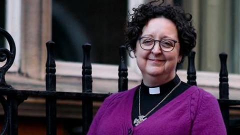 A woman standing on the pavement, she has black curly hair and is smiling while wearing a purple cardigan 
