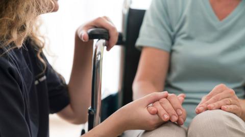 A close up of a woman in a wheelchair. She is wearing a light blue t-shirt and has her hands resting on her knees. A woman is squatting in front of her, holding onto the wheelchair rest with one hand while gently holding the woman's right hand.