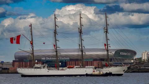 Landscape shot of the BAP Unión sailing on the River Mersey in Liverpool. The Hill Dickinson stadium is behind the ship. The Peru flag is hoisted up one of the ship's masts and is flying in the wind. 