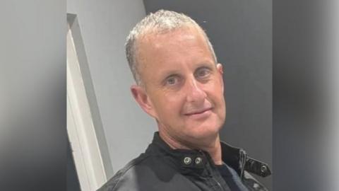 A man with grey hair wears a black jacket and navy T-shirt. He is looking at the camera.