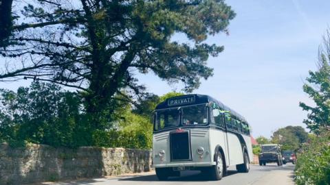 A vintage grey and black bus driving down a street in guernsey as part of the cavalcade